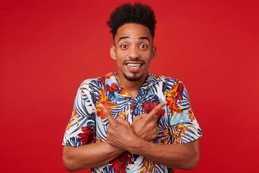 Portrait if young positive African American man in Hawaiian shirt, looks at the camera with cheerful expression, stands over red background and broadly smiles, points in different directions. photo