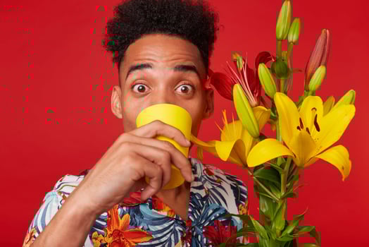 Close up of young African American man in Hawaiian shirt, surprised looks at the camera and drinking water from a yellow glass, holds yellow and red flowers bouquet, stands over red background. photo