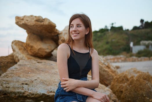 Photo of young cheerful ginger cute woman with freckles sits on the beach, sitting on a stone , dreamily looks away and smiles, looks positive and happy.