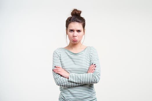 Offended young lovely brunette female with casual hairstyle crossing hands on her chest and looking sadly at camera with folded lips, standing over white background photo