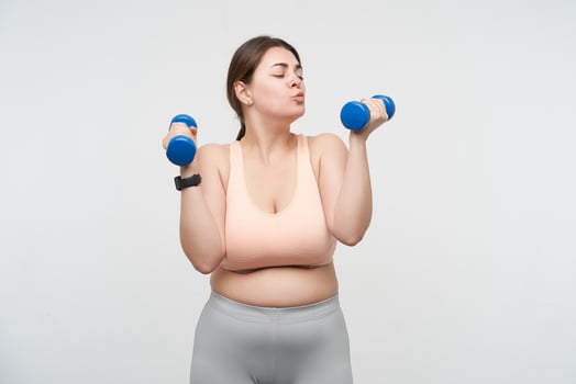 Positive young oversized female dressed in sports bra and leggins keeping blue dumbbells in raised hands and forming lips in kiss while looking at it, isolated over white background photo