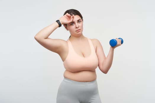 Exhausted young dark haired plump woman with blue dumbbells wiping sweat while being tired after hard training, posing over white background. Body positive concept photo