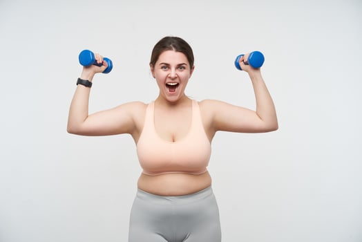 Excited young brown haired chubby female dressed in sporty top and leggins raising emotionally hands with dumbbells while posing over white background. Concept of self-made determination will-power photo