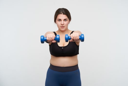 Concentrated young brunette chubby lady in sports bra and leggins keeping hands in front of herself while making exercises for hands, posing over white background photo