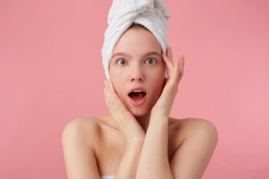 Close up of young amazed woman after shower with a towel on her head, looks at the camera with wide open mouth and eyes, touches face, stands over pink background. photo
