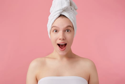 Close up of young happy amazed woman after shower with a towel on her head, looks at the camera with wide open mouth and eyes, hears cool news, stands over pink background. photo