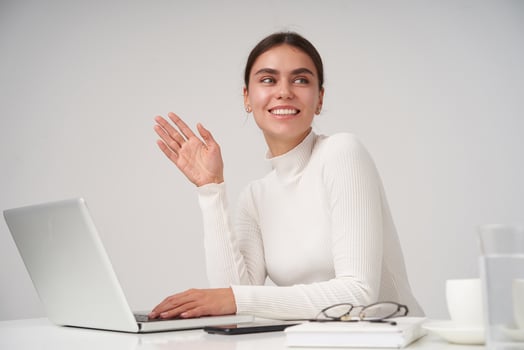 Cheerful young beautiful dark haired lady with natural makeup raising hand in hello gesture and looking positively aside with friendly smile, isolated over white background photo