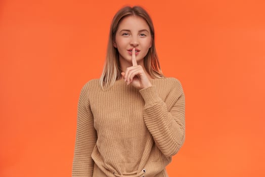 Studio photo of young blonde woman with short haircut keeping forefinger on her lips while asking to keep her secret and smiling cunningly, isolated over orange background