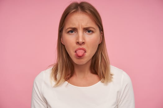Portrait of serious looking pretty blue-eyed female with blonde hair looking at camera with displeased face and showing tongue, standing over pink background in casual wear photo