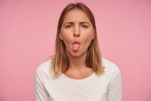 Portrait of beautiful blonde female with casual hairstyle frowning eyebrowns and showing her tongue while posing over pink background, wearing white blouse photo