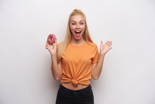 Overjoyed pretty young long haired blonde woman in casual clothes looking happily at camera with wide eyes and mouth opened, standing against white background with donut in raised hand photo