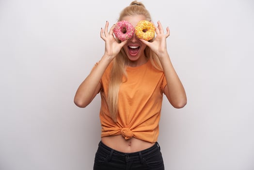 Joyous young long haired blonde slim woman fooling while standing against white background, looking at camera through colored donats and laughing cheerfully with wide mouth opened photo