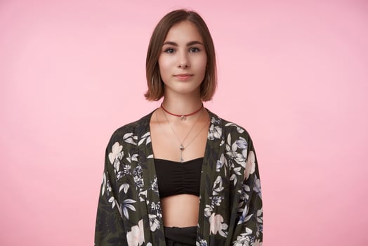 Indoor photo of young lovely short haired brunette female with natural makeup looking positively at camera with light smile while posing over pink background