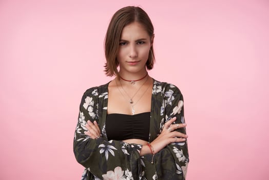 Attractive young brown-eyed brunette woman with natural makeup crossing her hands on chest while looking cunningly at camera, isolated over pink background photo