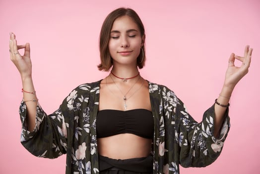 Pleasant looking young positive brunette woman raising hands in namaste gesture and smiling slightly while standing over pink background in flowered shirt photo
