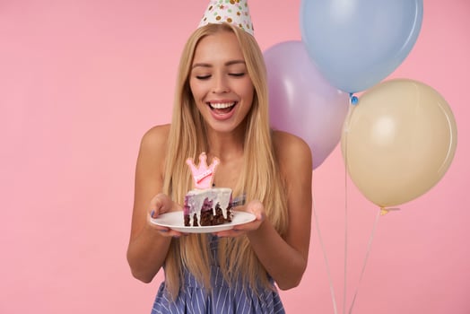 Portrait of cheerful young long haired woman wearing blue summer dress celebrating holiday, posing in multicolored air balloons with birthday cake, isolated over pink background photo