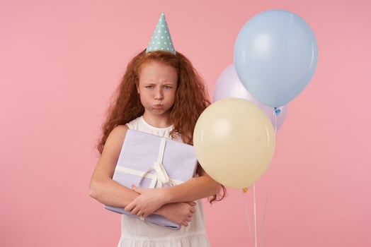 Offended little girl with long foxy hair holding wrapped present box, looking in camera sadly and pouting, isolated over pink background with colored air balloons, wearing white elegant dress photo