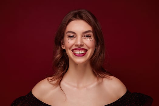 Close-up of charming young brown haired woman with wavy hairstyle wearing evening makeup with small stars on her face, smiling widely to camera while posing over claret background photo