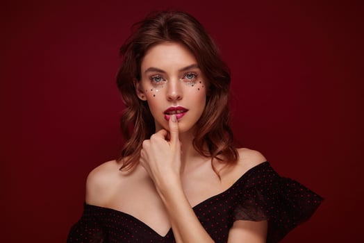 Pensive attractive young brunette female with festive hairdo and makeup looking at camera with calm face and keeping thumb on underlip, isolated over claret background in elegant black top photo