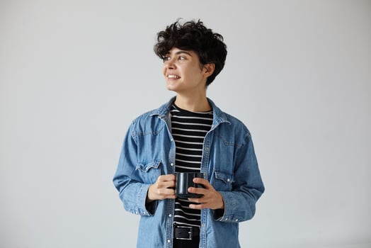 Stylish young pretty short haired curly brunette female holding cup of tea in raised hands and looking positively aside with pleasant smile, standing against white background photo