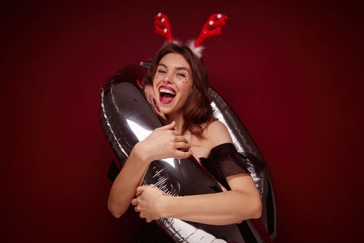 Studio shot of cheerful attractive young brown haired woman with evening makeup standing into number air balloon and laughing happily, wearing christmas horns while posing over claret background photo