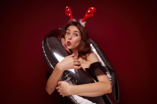 Funny shot of young pretty brunette lady wearing festive makeup while posing over claret background, enjoying x-mas theme party and posing with helium balloon photo