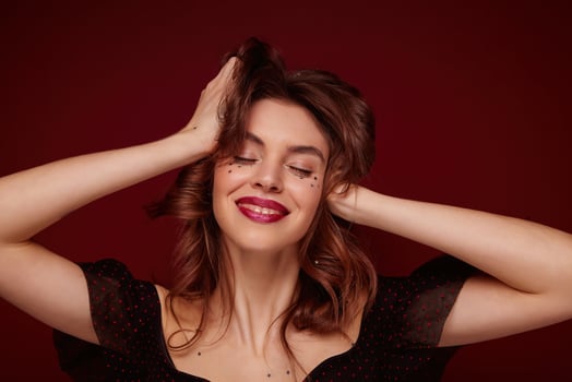 Pleased beautiful young brunette female with wavy hairstyle holding her head with raised hands and smiling positively, keeping eyes closed while standing over claret background photo