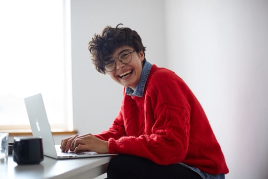 Joyful young dark haired curly lady with short trendy haircut holding her hands on keboerd of laptop while laughing happily, dressed in stylish casual clothes over white interior photo