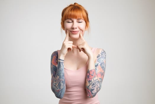 Pleasant looking young glad redhead tattooed lady with casual hairstyle keeping her eyes closed while smiling cheerfully, standing over white background photo