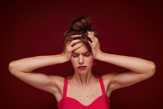 Displeased young brown haired female with bun hairstyle wearing pink top with straps while posing over burgundy background, frowning face and clutching her head, having headache after party photo