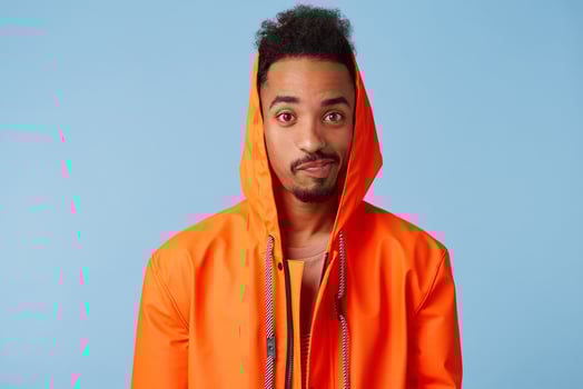 Unhappy handsome african american dark skinned boy wears in orange rain coat, confused, distressed by bad weather and spoiled weekend plans. Looks at the camera over blue background. photo