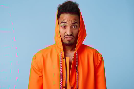Outraged african american guy in orange rain coat narrows his eyebrows and clenches his lips, looks at the camera very displeased, stands over blue background. photo