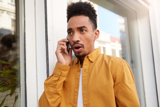 Young dark skinned amazed dark skinned man in yellow shirt, speaks on the phone with his friends and walking down the street, with shocked expression. photo