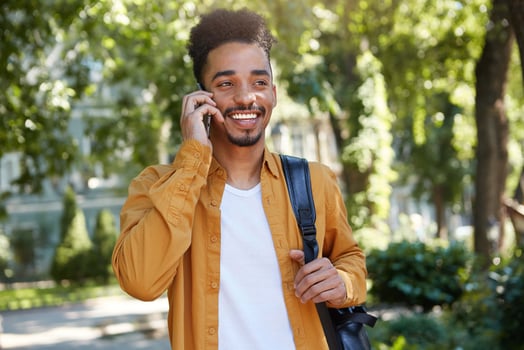 Young positive dark skinned man in yellow shirt, walking at the park, speaking on smartphone, waitng his friend, looking away and broadly smiling. photo