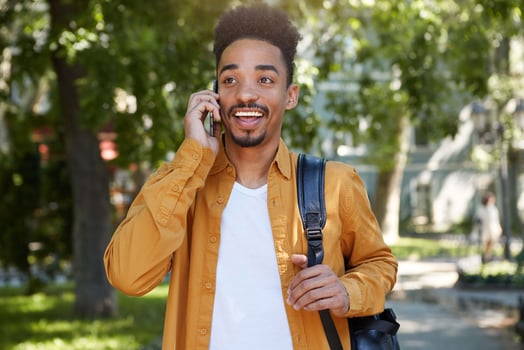 Photo of young smiling African American student boy in yellow shirt, walking at the park, speaking on smartphone with his friend, looking away and enjoy the day.