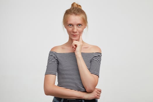 Pensive young green-eyed redhead female with casual hairstyle keeping her lips folded while looking at camera and holding forefinger on her cheek, posing over white background photo