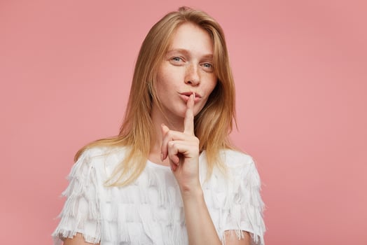 Positive young attractive female with foxy hair squinting her eyes and keeping raised forefinger on her mouth while posing over pink background, asking to be quiet photo