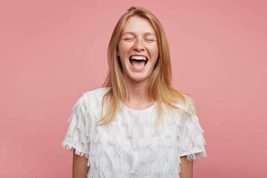 Good looking overjoyed young attractive female with foxy hair keeping her mouth wide opened while laughing happily with closed eyes, standing against pink background photo