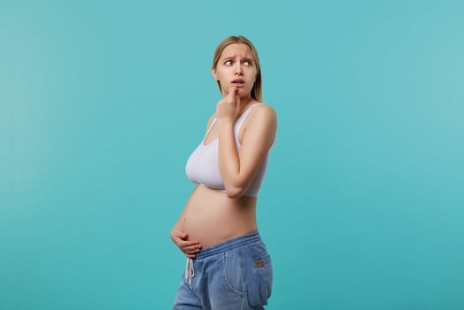 Frightened young blonde long haired woman being pregnant while standing over blue background, frowning eyebrows while looking scaredly aside and hugging her belly photo