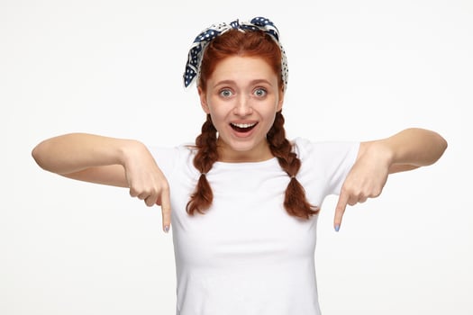 Indoor portrait of young ginger female posing over white background points with a finger to bottom side of the picture photo