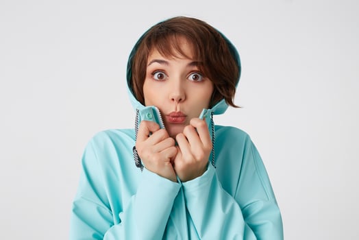 Photo of wondered young beautiful woman in blue rain coat, with a hood on his head, looks at the camera with surprise expressions, stands over white wall.