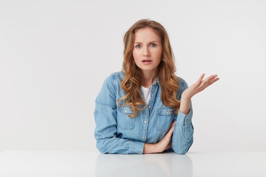 Portrait of confused young woman with long blond wavy hair, sitting at the table, one palm raised, looks skeptically displeased indignantly with incomprehension, over white background. photo