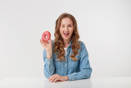 Portrait of cute young woman with long blond wavy hair, holds in his right hand a donut smiles and winks, isolated over white background. photo