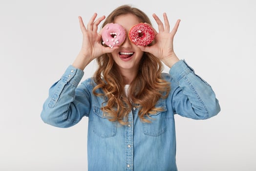 Young pretty woman with long blond wavy hair, wearing a denim shirt, looking through donuts and shows tongue and teeth, isolated over white background. photo