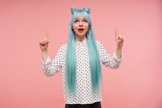 Positive young pretty woman with long blue anime hair showing upwards with raised index fingers while standing over pink background, wearing bright makeup and elegant clothes photo