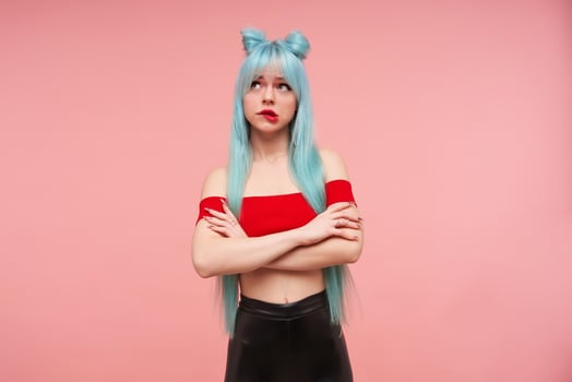Indoor shot of beautiful young woman with long blue anime hair crossing hands on her chest and looking thoughtfully upwards,biting underlip while posing over pink background in red top and black pants photo