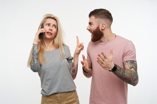 Stressed young pretty brown haired bearded man looking angrily on his attractive blonde tattooed girlfriend while she is having phone conversation, isolated over white background photo