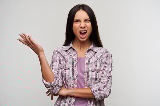 Indignant young dark haired lady with natural makeup raising palm emotionally while standing over white background, looking angily to camera and frowning her face photo