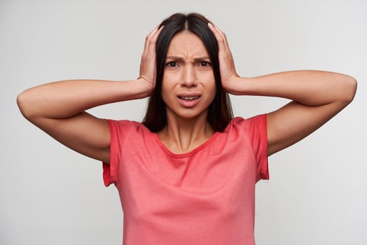 Displeased young attractive brunette woman with casual hairstyle covering her ears and frowning her face, trying to avoid annoying sounds, dressed in pink t-shirt over white background photo
