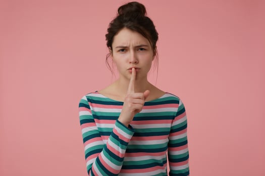 Young lady, pretty woman with brunette hair and bun. Wearing striped blouse and showing sign of silence, touching lips with a finger. Watching at the camera isolated over pastel pink background photo